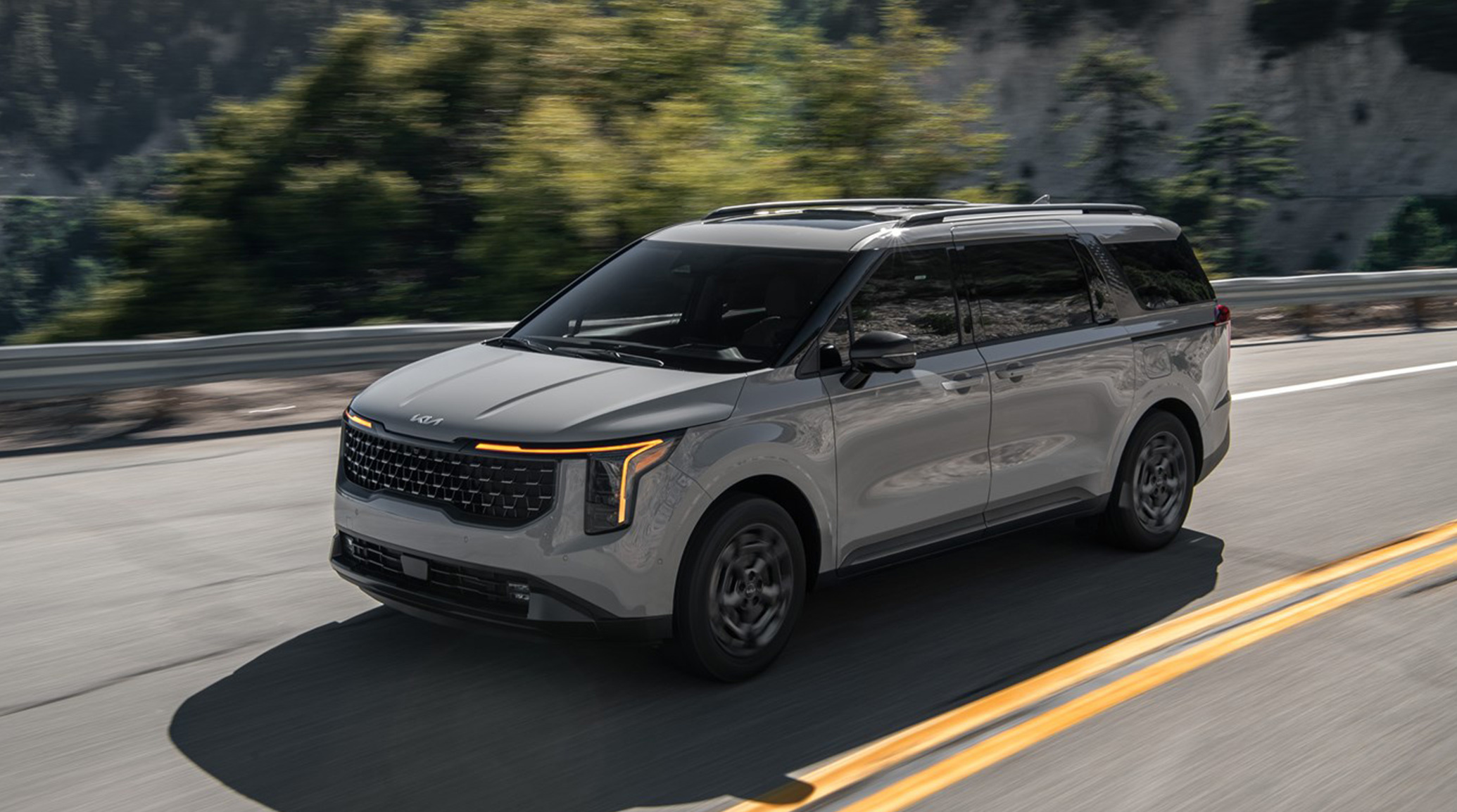 2025 Kia Carnival in gray, driving on an open road on a hill-side highway, front three-quarter view