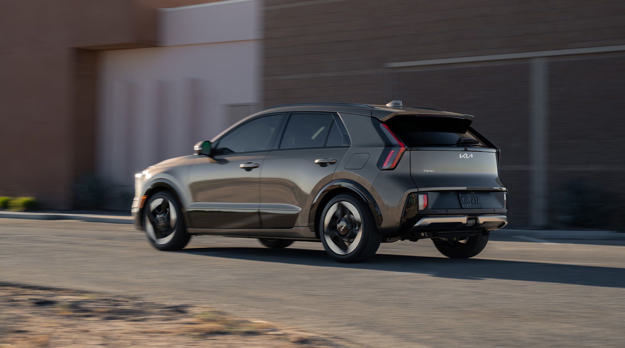 Rear angle photograph of the 2027 Kia Niro Hybrid Electric SUV driving over an outdoor road with a building behind it.