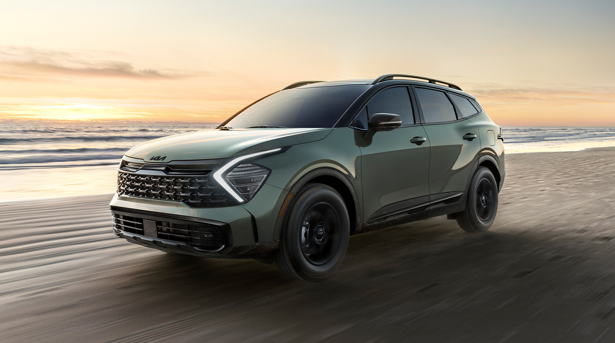 2025 Kia Sportage in green, in a three-quarter driver side view, driving down a beach near the water