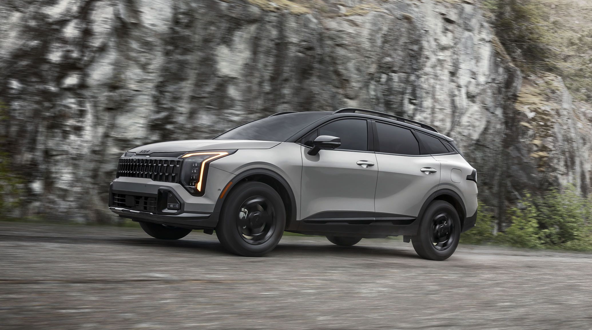 The 2027 Kia Sportage SUV in grey with glowing orange headlights, driving boldly in front of a motion-blurred rocky cliff.