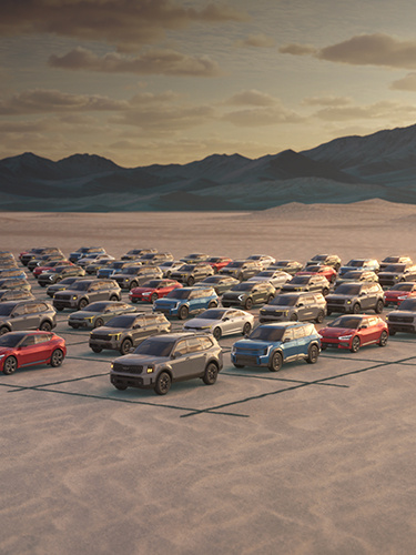 A large group of Kia vehicles in a variety of colors and models parked on a flat, open area with mountains in the background.