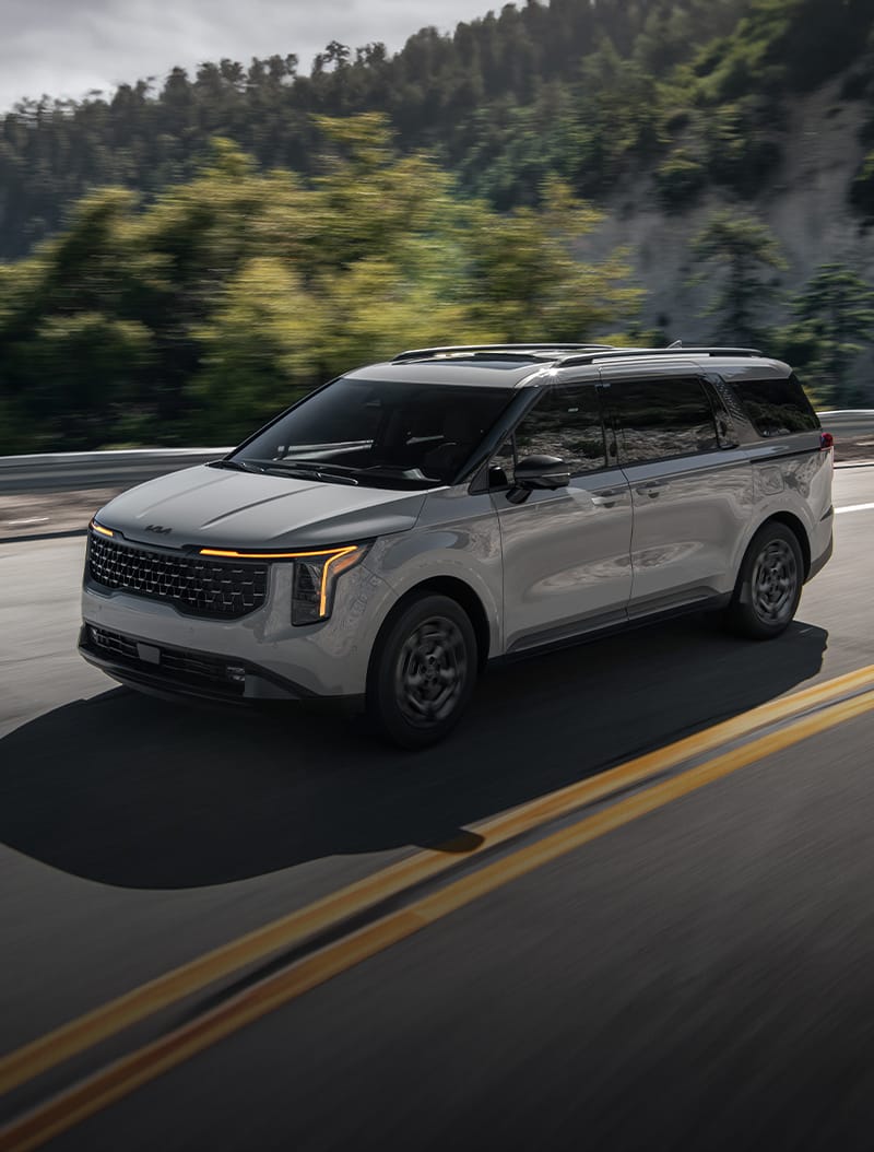 The Kia Carnival MPV Hybrid in gray, highlighting the exterior design and bold grille. The SUV-inspired minivan is driving quickly on a road with mountains and greenery in the backdrop.
