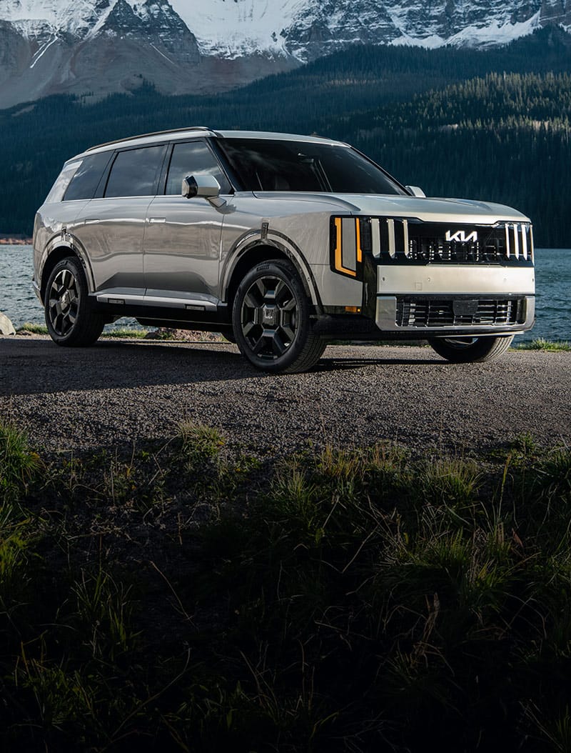 A silver 2027 Kia Telluride Hybrid parked on a rocky lakeside with towering snow-covered mountains in the background, highlighting the SUV’s bold exterior design and rugged capabilities.