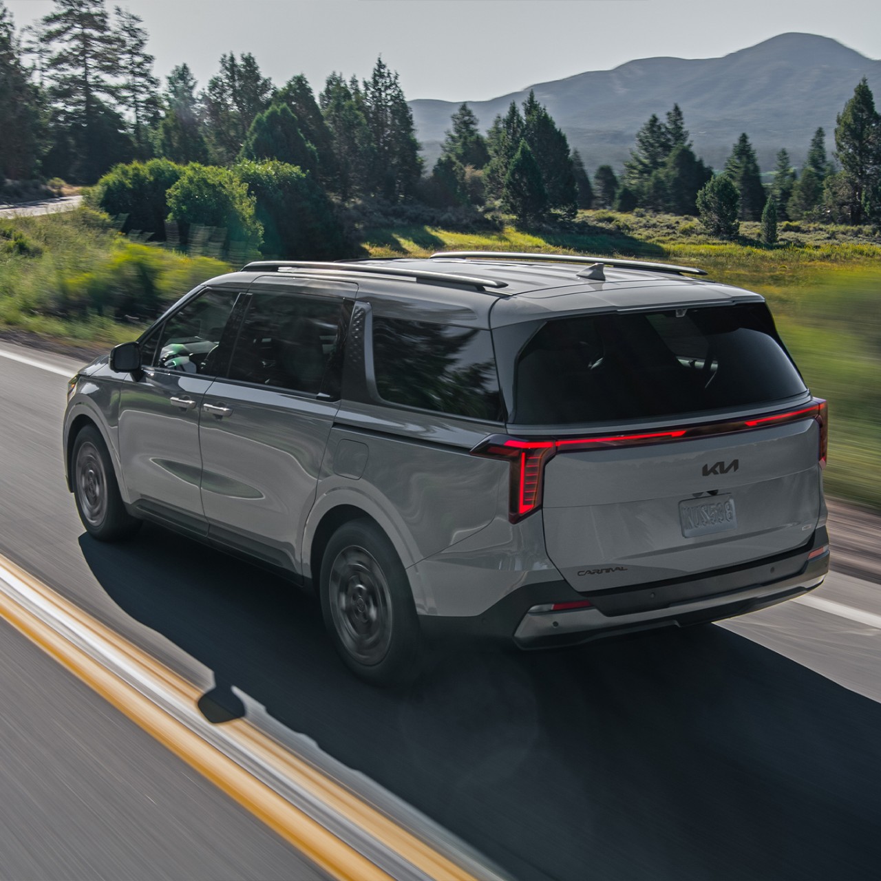 2025 Kia Carnival MPV Hybrid, three-quarter back view, driving on a road by lush trees featuring vertical LED tail lights